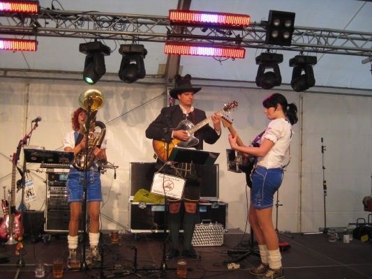 Eine Band spielt auf einer Bühne mit einem Mann, der Gitarre spielt, einer Frau, die Gitarre spielt, und einer Frau, die Tuba spielt. Sie stehen unter Bühnenbeleuchtung mit einem weißen Hintergrund.