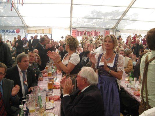 Eine große Gruppe von Menschen versammelt sich in einem Zelt, klatscht und jubelt. Ein Mann in Anzug sitzt an einem Tisch mit einer Frau in traditioneller Kleidung. Eine Band spielt im Hintergrund.