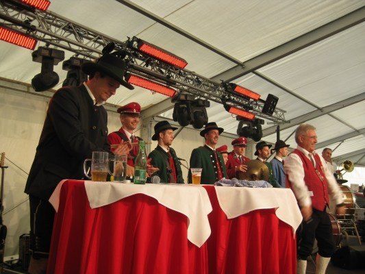 Eine Gruppe von Männern in traditioneller bayerischer Kleidung, einschließlich Hüten und Westen, steht um einen Tisch mit Bierkrügen und Flaschen, möglicherweise bei einem Festival. Ein Mann spricht in ein Mikrofon.