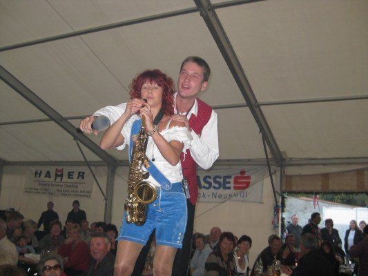 Eine Frau in traditioneller bayerischer Kleidung spielt Saxophon, während ein Mann hinter ihr steht, beide treten auf einer Bühne unter einem Zelt für ein Publikum auf.