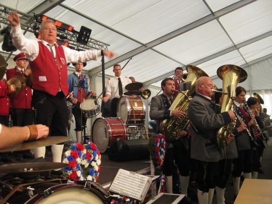 Ein Blasorchester spielt auf der Bühne, mit Musikern, die Tubas, Trommeln und Becken spielen. Der Dirigent, der eine rote Weste trägt, führt das Ensemble an. Die Bühne ist unter einem weißen Zelt aufgebaut.