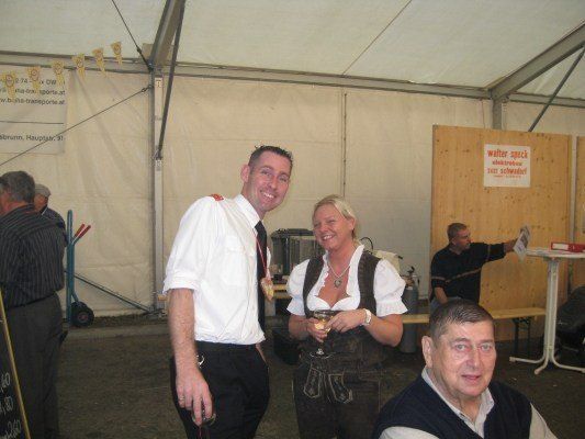 Ein Mann und eine Frau in traditioneller bayerischer Kleidung lächeln und halten Weingläser in einem Zelt. Dahinter steht ein Mann in der Nähe einer Holzwand mit einem Schild.