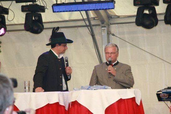 Zwei Männer stehen mit Mikrofonen an einem Podium, einer trägt einen traditionellen Hut, unter Bühnenbeleuchtung.
