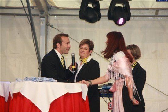 Ein Mann und eine Frau in formeller Kleidung schütteln sich die Hände bei einer Veranstaltung. Eine Frau in einem rosa Kleid steht neben ihnen. Ein Mikrofon wird von einem der Männer gehalten.