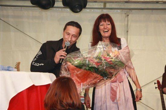 Ein Mann mit Mikrofon und Blumenstrauß steht neben einer lächelnden Frau in traditioneller Kleidung.