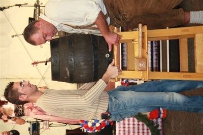 Zwei Männer arbeiten gemeinsam daran, einen großen Holzfass mit einem Holzwerkzeug anzuzapfen. Ein Mann trägt eine Brille und ein gestreiftes Hemd, während der andere in einer braunen Schürze ist.