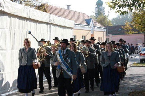 Eine Musikkapelle in traditioneller Kleidung, einschließlich Männer und Frauen, geht eine Straße hinunter, mit Gebäuden und Bäumen im Hintergrund.