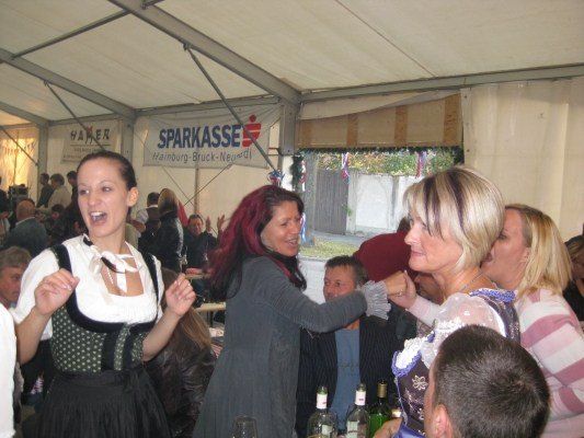 Eine Gruppe von Menschen versammelt sich in einem Zelt, einige tragen traditionelle Kleidung. Eine Frau mit roten Haaren hält eine Flasche und scheint mit einer anderen Frau zu sprechen. Ein Banner mit dem Text 'SPARKASSE' ist im Hintergrund sichtbar.