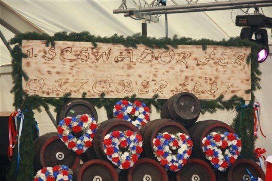 Ein Holzschild mit '1. SCHWADORFER OCTOBERFEST' darauf, über einem Stapel Fässer, geschmückt mit roten, weißen und blauen Kränzen.