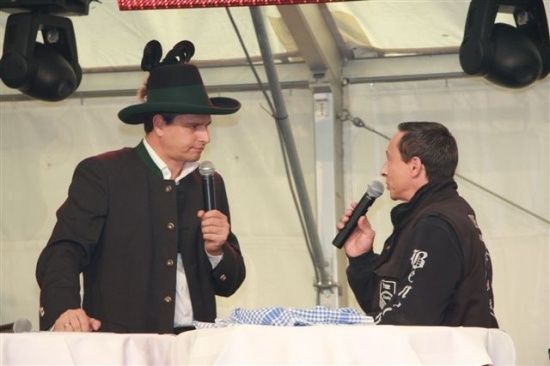 Zwei Männer stehen an einem Podium, einer trägt einen traditionellen bayerischen Hut und der andere ein schwarzes Jackett. Beide halten Mikrofone und scheinen zu sprechen.