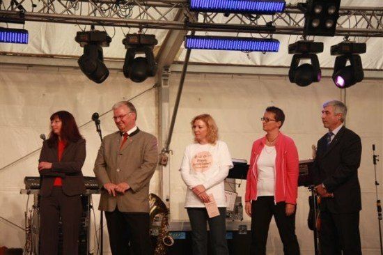 Fünf Personen stehen auf einer Bühne unter einem Zelt, mit Musikinstrumenten und Mikrofonen im Hintergrund.