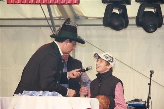 Ein Mann in einem Hut hält ein Mikrofon und spricht zu einem jungen Jungen, der eine Baseballkappe und eine Weste trägt. Sie stehen vor einem Tisch.