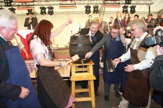 Eine Gruppe von Menschen in Schürzen klopft mit einem Hammer auf einen Fass in einer festlichen Umgebung, während eine Menge zuschaut.