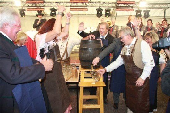 Eine Gruppe von Menschen in traditioneller Kleidung versammelt sich um einen großen Fass, wobei ein Mann Bier in ein Glas gießt. Die Szene scheint eine Feier oder ein Festival zu sein.
