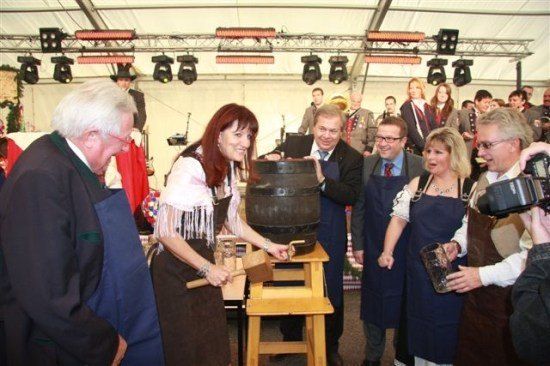 Eine Gruppe von Menschen in traditioneller Kleidung versammelt sich um einen großen Holzfass, eine Frau hält einen Hammer und ein Mann ein Glas. Sie befinden sich in einer festlichen Umgebung mit Bühnenbeleuchtung und einer Menge.