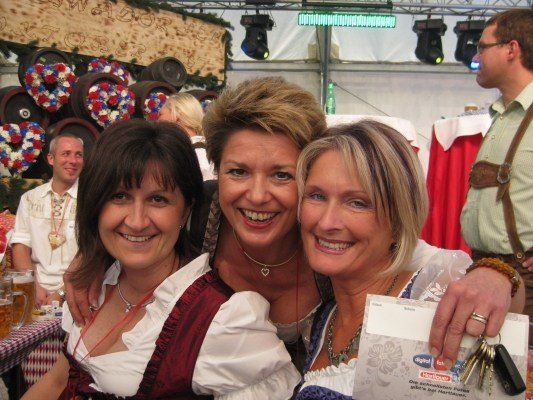 Drei Frauen in traditioneller bayerischer Kleidung lächeln und posieren für ein Foto bei einer festlichen Veranstaltung, eine hält einen Schlüssel und ein Papier. Im Hintergrund befinden sich Fässer, ein Tisch mit Bierkrügen und ein Mann in einem grünen Hemd.