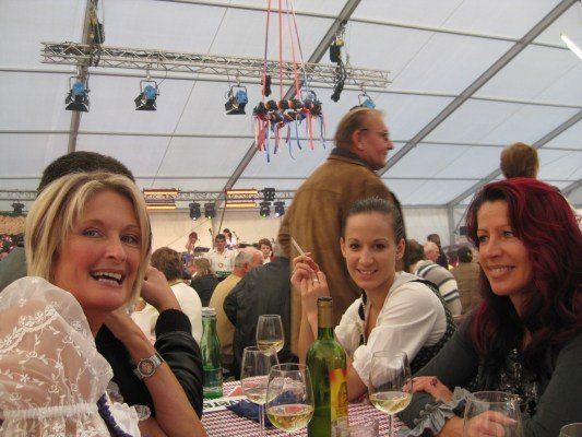 Drei Frauen sitzen in einem Zelt an einem Tisch, lächeln und halten Weingläser. Eine Frau raucht eine Zigarette. Ein Mann geht hinter ihnen, und Bühnenlichter hängen von der Decke.
