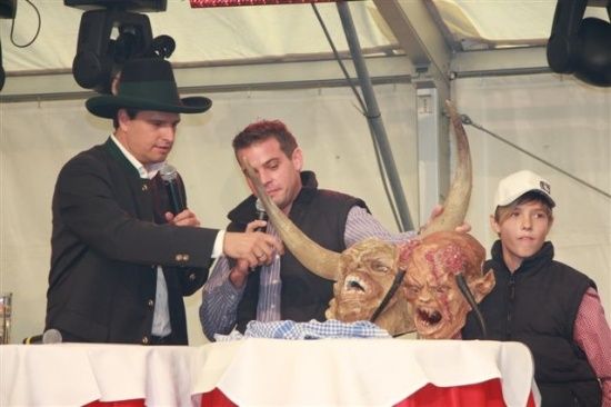 Drei Männer stehen an einem Tisch mit einer großen gehörnten Maske. Der Mann auf der linken Seite hält ein Mikrofon und spricht, während der Mann in der Mitte den Horn der Maske hält. Der Mann auf der rechten Seite trägt einen Hut und schaut auf die Maske.
