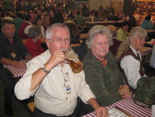 Ein älterer Mann trinkt aus einem großen Bierkrug, während er mit zwei älteren Frauen an einem Tisch in einem überfüllten Raum sitzt.