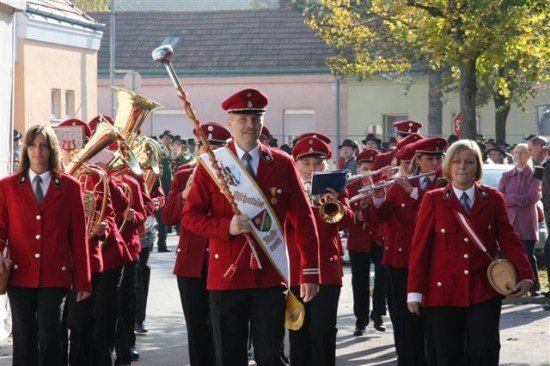 Ein Musikzug in roten Uniformen, angeführt von einem Mann mit einem Dirigentenstab, tritt auf einer Straße auf, mit Zuschauern.