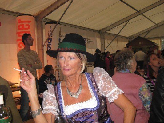 Eine Frau in traditioneller bayerischer Kleidung raucht eine Zigarette bei einer Veranstaltung im Freien. Sie trägt einen Hut und eine Halskette. Hinter ihr versammeln sich mehrere Personen, einige stehend und andere sitzend. Ein Mann in einem gestreiften Shirt steht im Hintergrund.