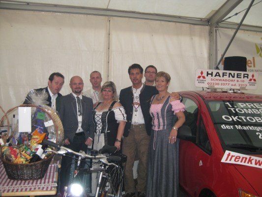 Eine Gruppe von Menschen in traditioneller Kleidung posiert für ein Foto vor einem roten Auto und einem Fahrrad. Das Auto hat ein Schild mit der Aufschrift 'Hafner' und ein Korb mit Gegenständen steht auf dem Tisch.
