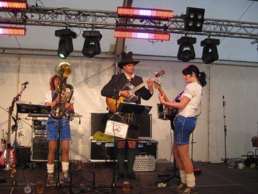 Eine Band spielt auf der Bühne, ein Mann spielt Gitarre, eine Frau spielt Saxophon und eine weitere Frau spielt Gitarre. Die Bühne ist mit Mikrofonen, Lautsprechern und Beleuchtung ausgestattet.