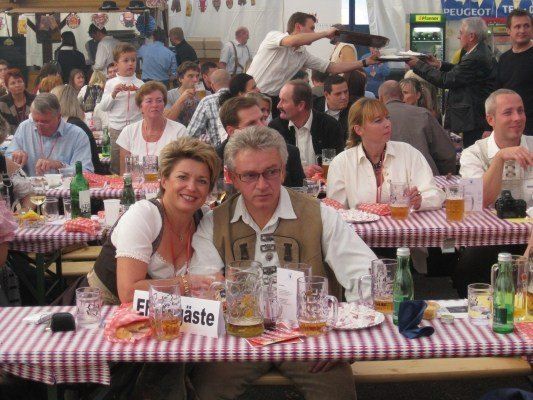 Eine Gruppe von Menschen sitzt in einem Biergarten an einem Tisch und lächelt. Der Tisch ist mit einer rot-weiß karierten Tischdecke bedeckt. Der Mann und die Frau vor ihnen tragen eine Brille und haben ein Schild mit der Aufschrift 'El Paste'. Es gibt andere Leute, die an anderen Tischen sitzen, und ein Mann steht hinter ihnen.