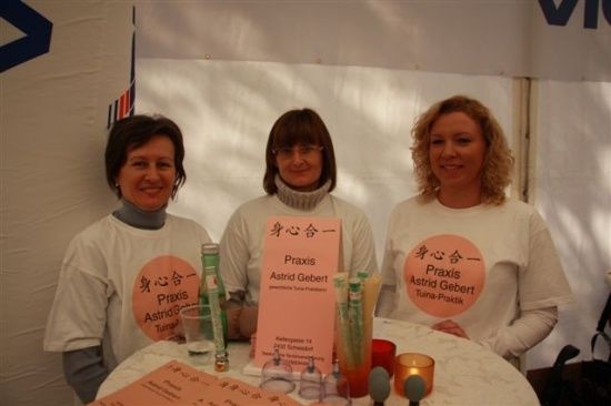 Drei Frauen in weißen T-Shirts mit rosa Logos stehen hinter einem Tisch mit Tassen und Gläsern. Sie lächeln und scheinen für ein Foto zu posieren.