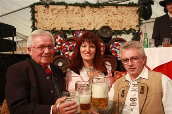 Drei Personen, zwei Männer und eine Frau, lächeln und halten große Bierkrüge hoch in einer festlichen Umgebung mit Fässern und einem dekorierten Hintergrund.