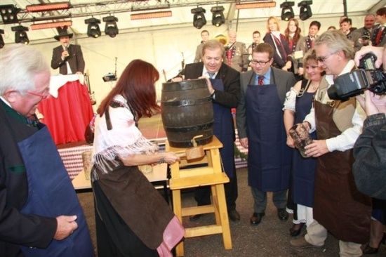 Eine Frau klopft mit einem Hammer auf einen großen Holzfass, während ein Mann in einem Anzug zusieht. Mehrere Personen in Schürzen umgeben sie, und ein Mann mit einer Kamera hält den Moment fest. Im Hintergrund spielt ein Musiker ein Blechinstrument.