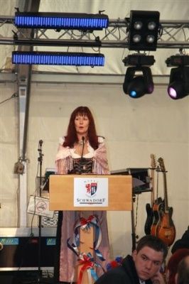 Eine Frau steht an einem Podium mit einem Mikrofon, trägt ein Kostüm und ist von Musikinstrumenten und Bühnenbeleuchtung umgeben.