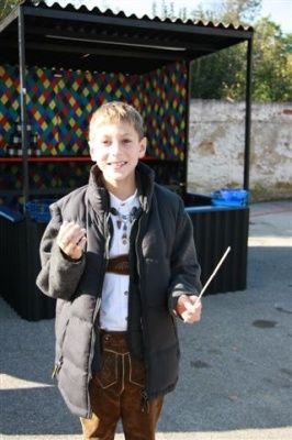 Ein junger Junge in traditioneller bayerischer Kleidung steht draußen, hält einen Stock in der Hand und lächelt für die Kamera. Hinter ihm befindet sich ein farbenfroher Stand mit verschiedenen ausgestellten Artikeln.