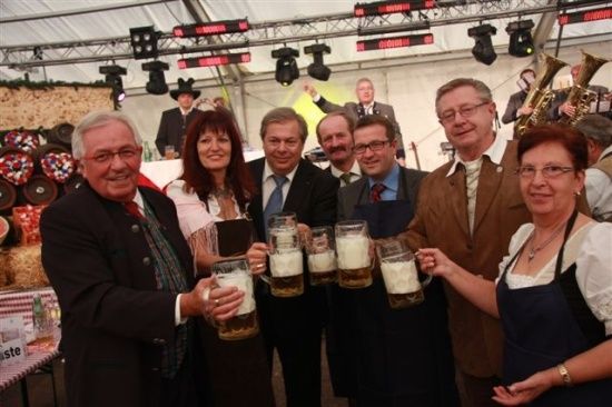 Eine Gruppe von Menschen in formeller Kleidung hält Bierkrüge in einer festlichen Umgebung hoch, während eine Band im Hintergrund spielt.