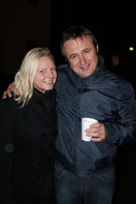Ein Mann und eine Frau stehen bei Nacht eng beieinander und lächeln für die Kamera. Der Mann hält einen weißen Becher in der Hand.