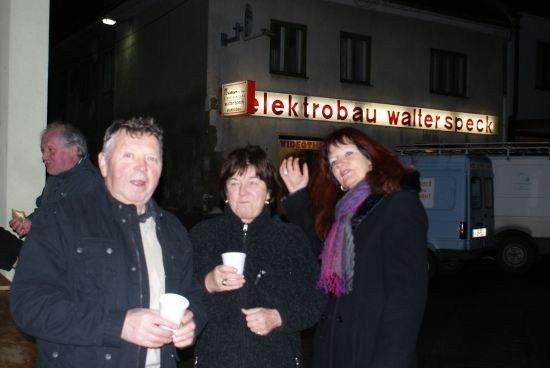Drei Menschen stehen nachts vor einem Gebäude, einer hält einen Becher, ein anderer winkt und ein Lieferwagen ist geparkt.