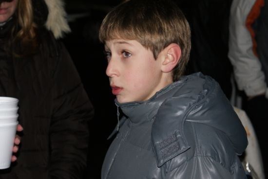 Ein junger Junge in einem grauen Mantel steht in einem schwach beleuchteten Bereich und schaut zur Seite. Hinter ihm hält eine Person einen Stapel weißer Becher.