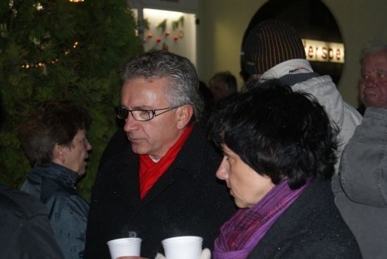 Ein Mann und eine Frau, beide mit weißen Bechern in der Hand, stehen eng beieinander in einer Menschenmenge. Der Mann trägt eine Brille und ein rotes Halstuch, während die Frau ein lila Halstuch hat. Hinter ihnen ist ein Weihnachtsbaum zu sehen.