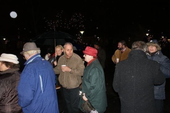 Eine Gruppe von Menschen steht bei Nacht draußen, einige tragen Hüte und halten Tassen. Sie werden von Straßenlaternen und Feuerwerk im Hintergrund beleuchtet.