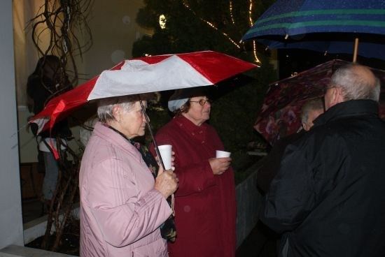 Zwei Frauen in Mänteln und Mützen, eine hält einen Becher, stehen unter einem Regenschirm bei Regen. Ein Mann in einem schwarzen Mantel steht in der Nähe.