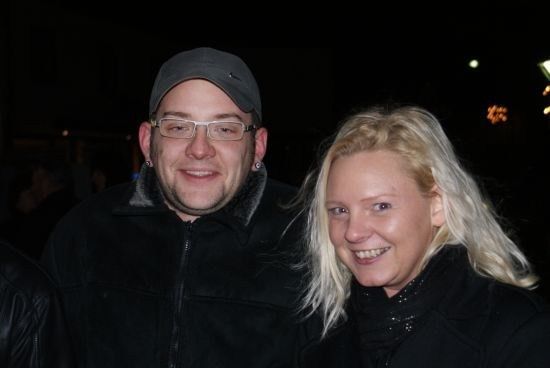 Ein Mann und eine Frau lächeln für ein Foto bei Nacht, beide tragen schwarze Jacken. Der Mann hat eine Brille und eine Kappe.