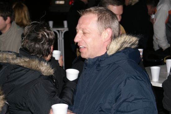 Ein Mann in einer blauen Jacke mit einem Pelzkragen lächelt, während er einen Becher hält, umgeben von Menschen bei einer Zusammenkunft.