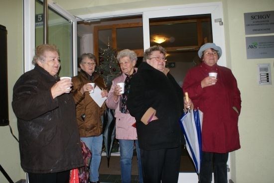 Fünf Frauen in Wintermänteln stehen in einer Tür, halten Tassen und Papiere in der Hand, mit einem Weihnachtsbaum im Hintergrund.