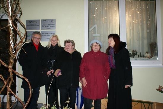 Fünf Menschen stehen vor einem Gebäude, lächeln und posieren für ein Foto. Sie tragen alle Winterkleidung. Hinter ihnen befindet sich ein Fenster mit einem weißen Vorhang und einem Schild.