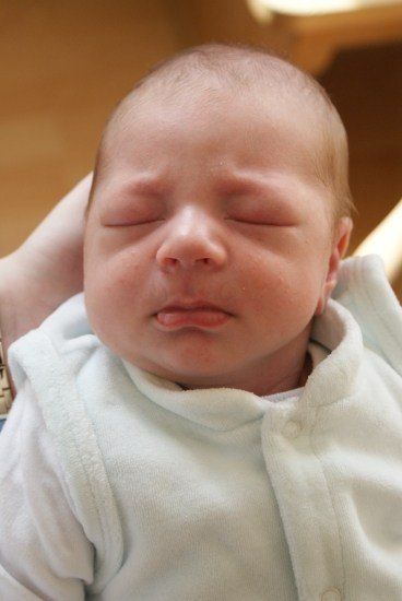 Ein Neugeborenes mit geschlossenen Augen, leicht geöffnetem Mund und in einer weißen Kleidung wird von jemandem gehalten.
