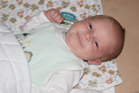 Ein lächelndes Baby liegt auf einer Decke mit einem bärengemusterten Tuch und einem Rasselspielzeug in der Hand. Das Baby trägt ein grünes Lätzchen mit Schmetterlingsdesign.