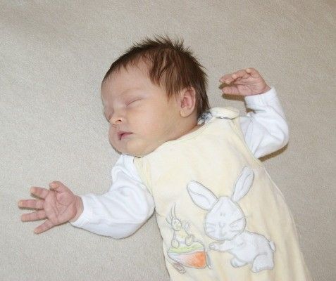Ein schlafendes Baby in einem gelben Hasen-Overall liegt auf einer beigen Oberfläche, mit beiden Händen erhoben.