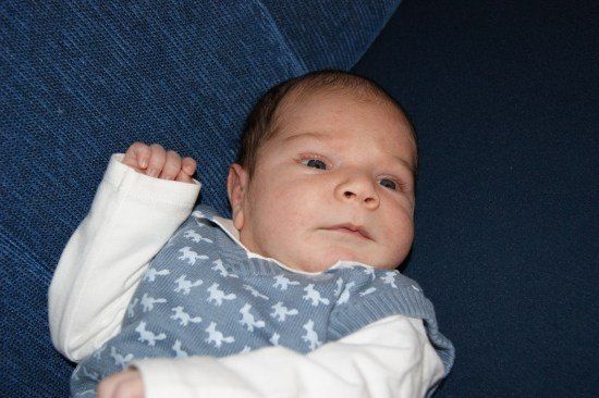 Ein Baby liegt auf einer blauen Couch und trägt ein blau-weiß gemustertes Outfit, während es neugierig nach oben schaut.