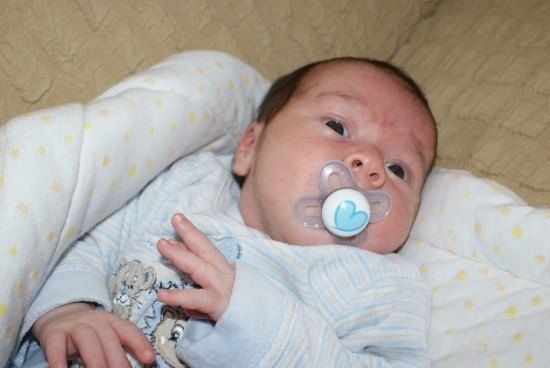 Ein Baby mit einem Schnuller im Mund, auf einer Decke mit gelbem Sternenmuster liegend. Das Baby trägt ein blau-weiß gestreiftes Outfit.