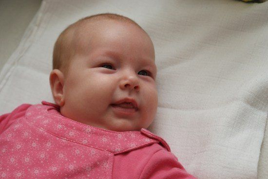 Ein Babymädchen liegt auf einer weißen Decke und trägt ein rosa Shirt mit weißem Schneeflockenmuster. Sie hat ein leichtes Lächeln auf dem Gesicht.
